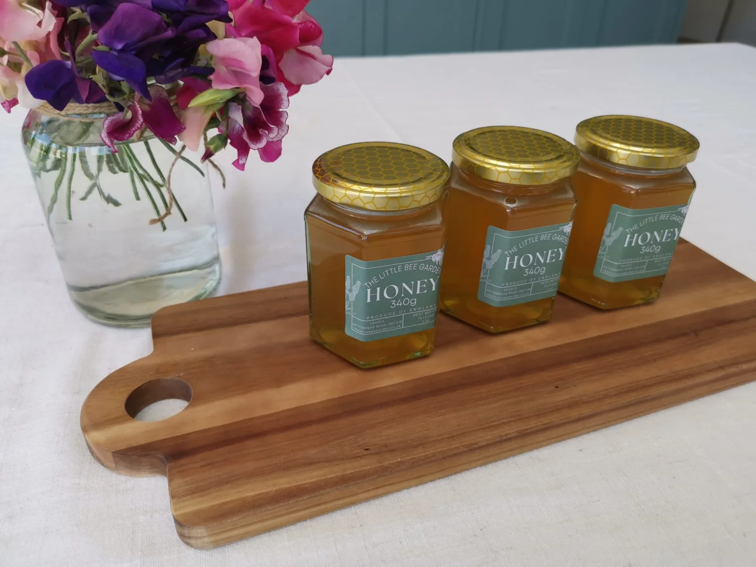Three jars of The Little Bee Garden honey on a wooden board beside fresh flowers